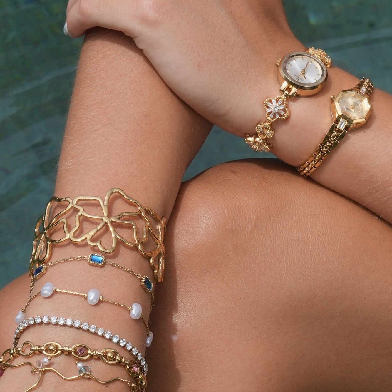Nahaufnahme des Arms einer Person mit mehreren goldenen Armbändern und Ringen vor einem unscharfen Naturhintergrund